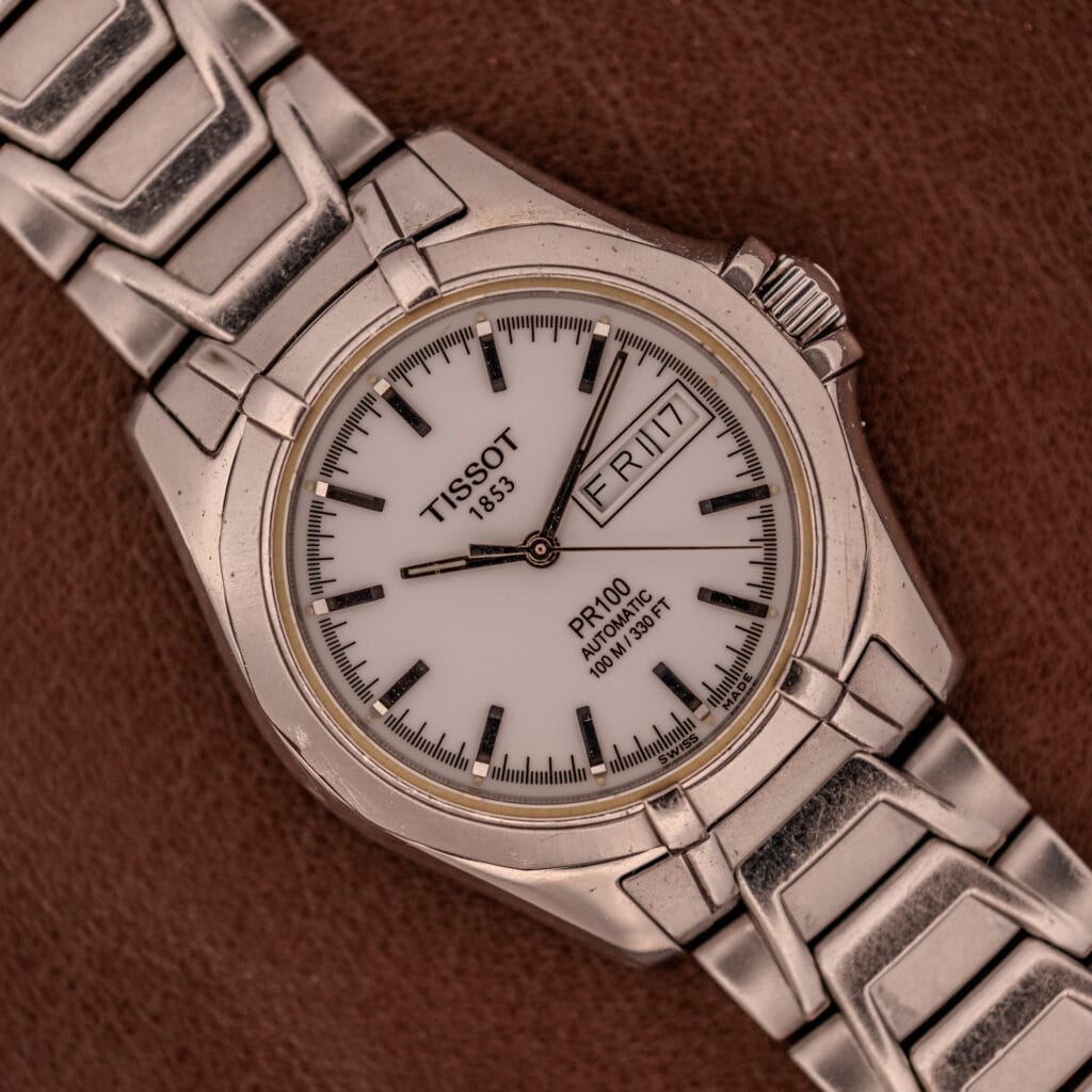 The Tissot PR100 Automatic Day/Date P 764 in stainless steel, featuring a white dial, is displayed against a brown background.