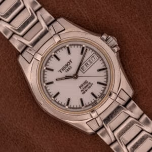 Tissot PR100 Automatic Day/Date Ref. P 764 shown flat on brown leather with the steel bracelet folded back behind the matte-white dial.