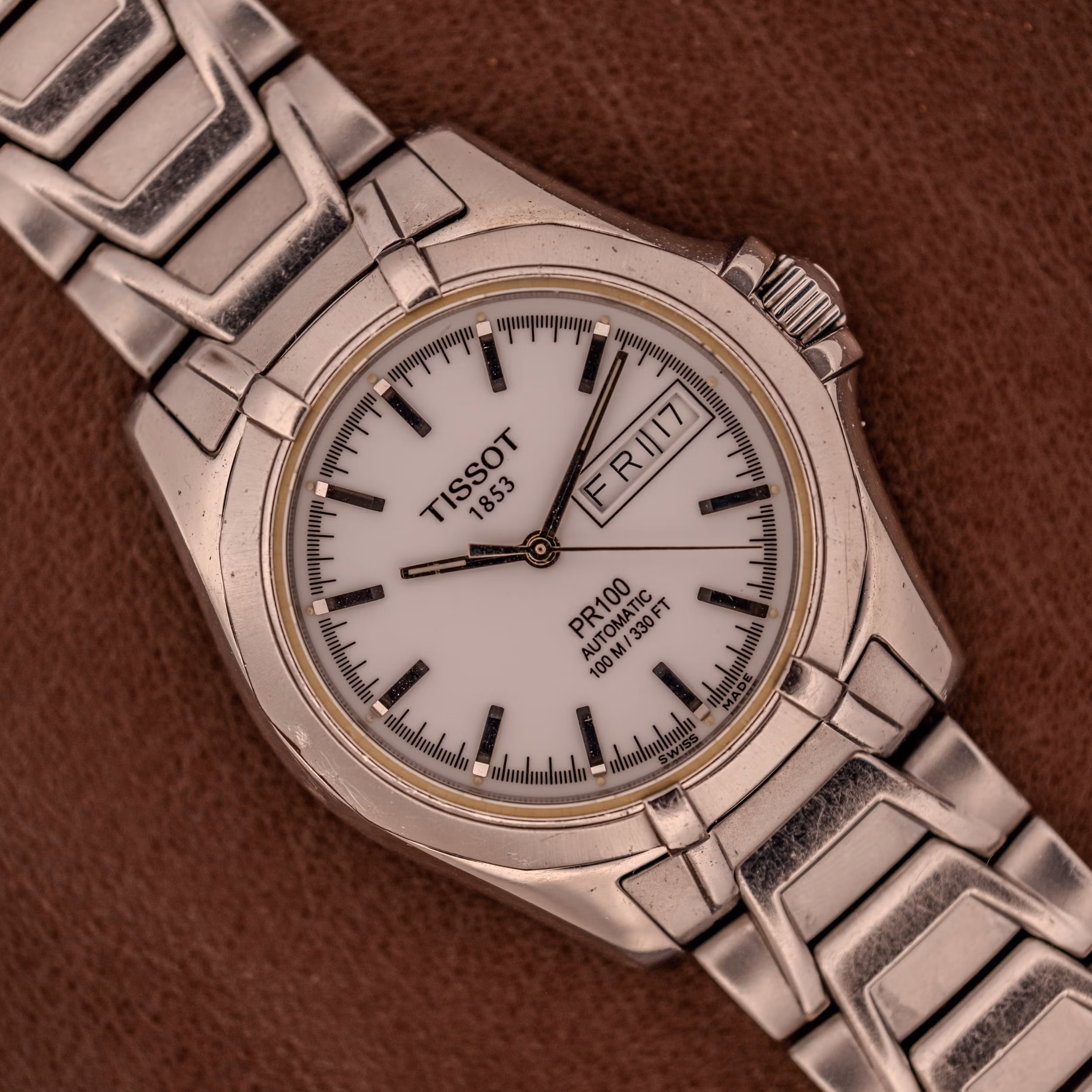 The Tissot PR100 Automatic Day/Date P 764 in stainless steel, featuring a white dial, is displayed against a brown background. Tissot PR100 Automatic Day/Date Ref. P 764 shown flat on brown leather with the steel bracelet folded back behind the matte-white dial.