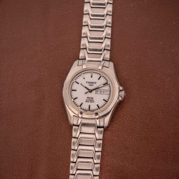 A Tissot PR100 Automatic Day/Date P 764 wristwatch in stainless steel is shown with a sleek silver design, resting on a brown textured surface.