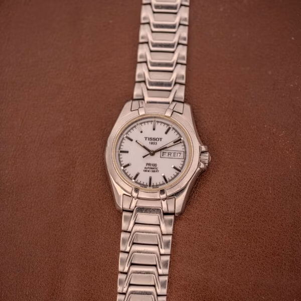 Angled view of the Tissot PR100 Automatic showing the polished sloped bezel, integrated steel bracelet links, and signed crown at three.