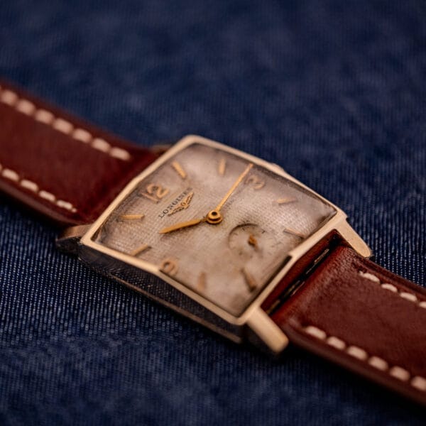 A Longines Square 10k Gold Filled Case Waffle Dial Cal. 25Z wristwatch with a brown leather strap rests on blue denim fabric.
