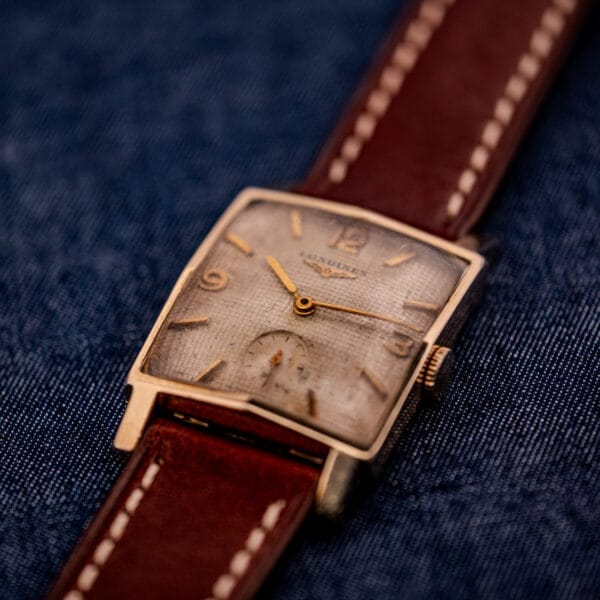 A Longines Square 10k Gold Filled Case Waffle Dial Cal. 25Z wristwatch with a brown leather strap rests on blue denim fabric.