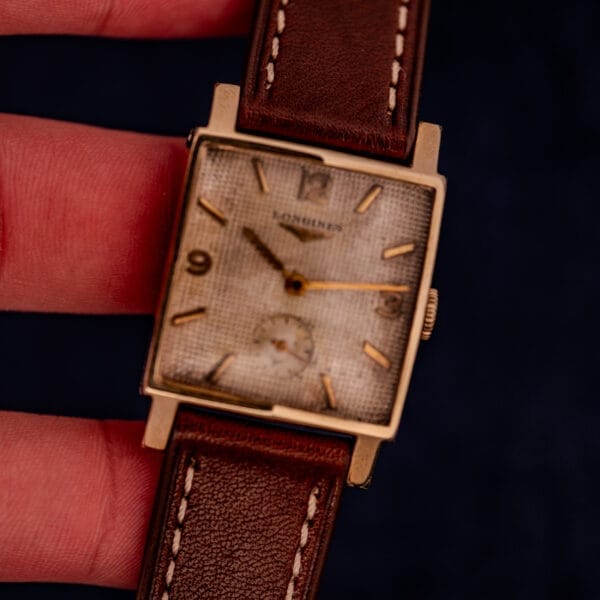 A hand holds a Longines Square 10k Gold Filled Case watch with a waffle dial (Cal. 25Z) and a brown leather strap.