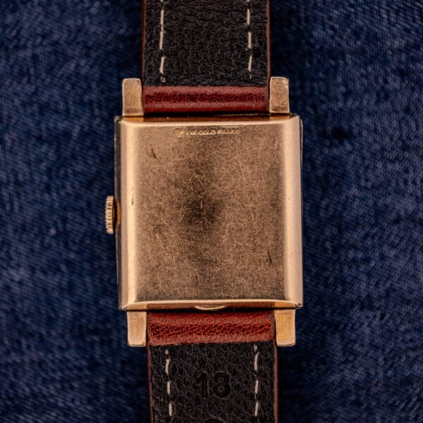 A Longines Square 10k Gold Filled Case Waffle Dial Cal. 25Z wristwatch with a black leather strap is displayed on a blue fabric background.