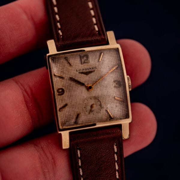 A hand holds a Longines Square 10k Gold Filled Case Waffle Dial Cal. 25Z wristwatch with a brown leather strap and distinctive waffle dial.
