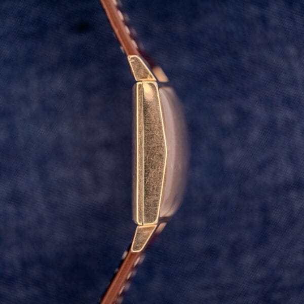 Side view of a Longines Square 10k Gold Filled Case Waffle Dial Cal. 25Z wristwatch with a brown leather strap, set against a blue textured background.