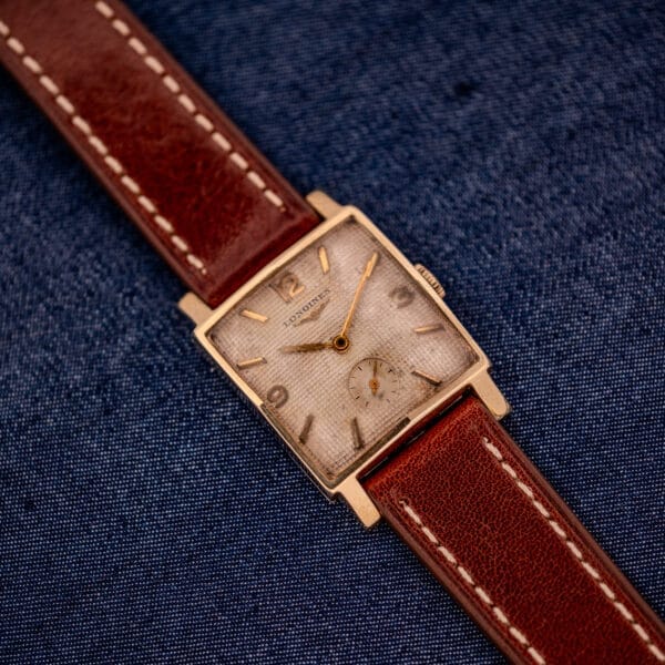 A vintage Longines Square 10k Gold Filled Case Waffle Dial Cal. 25Z wristwatch with a brown leather strap, displayed on blue denim fabric.