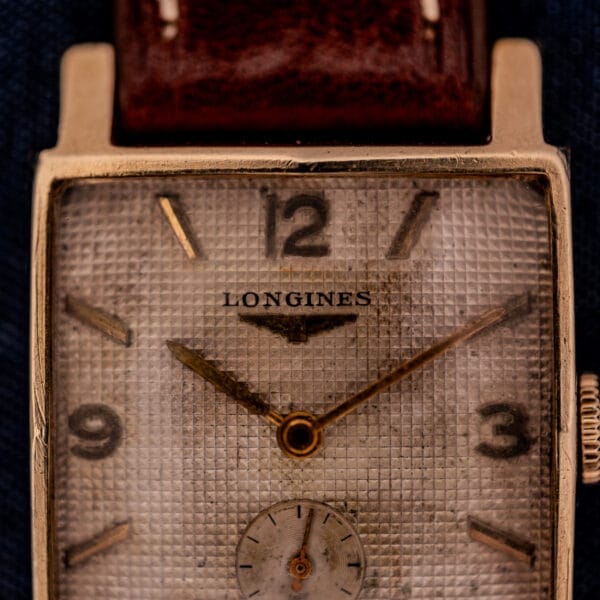 Close-up of a vintage Longines Square 10k Gold Filled Case Waffle Dial Cal. 25Z wristwatch, featuring a worn textured waffle dial and brown leather strap.