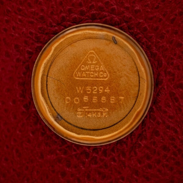 The Omega Ladymatic Sunburst Dial Ref. W 5294 Cal. 680 gold case back rests on a red textured background, engraved with OMEGA WATCH CO, Cal. 680 numbers, and hallmark.