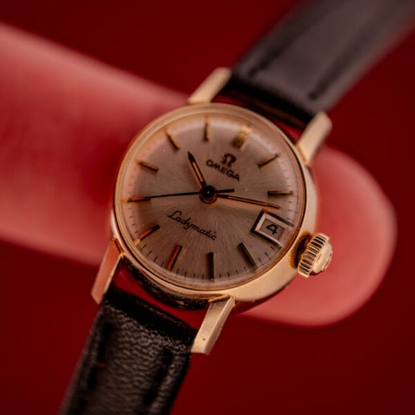 Close-up of an Omega Ladymatic Sunburst Dial Ref. W 5294 Cal. 680 with a black strap and gold casing, displayed on a red background.