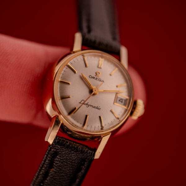 A close-up of the Omega Ladymatic Sunburst Dial Ref. W 5294 Cal. 680, highlighting its gold case, striking sunburst dial, black leather strap, and date display.