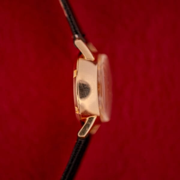 Omega Ladymatic Sunburst Dial Ref. W 5294 Cal. 680 gold wristwatch with a black strap, shown from the side against a red textured background.