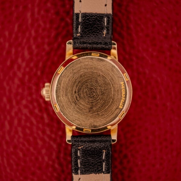 The Omega Ladymatic Sunburst Dial Ref. W 5294 Cal. 680 shines with a gold finish, black leather strap, and a circular back displaying its refined movement on a textured red surface.