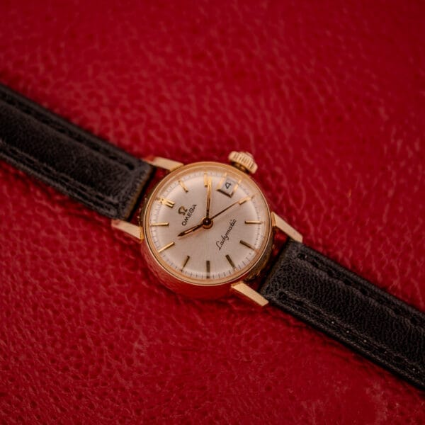 An Omega Ladymatic Sunburst Dial Ref. W 5294 Cal. 680 wristwatch with a gold case and black leather strap is displayed on a textured red background.