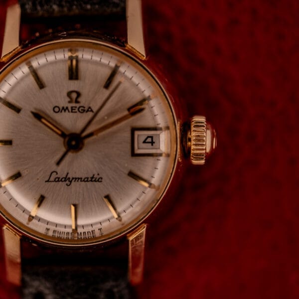 Close-up of the Omega Ladymatic Sunburst Dial Ref. W 5294 Cal. 680 gold wristwatch with date display, set against a red background.