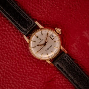 An Omega Ladymatic Sunburst Dial Ref. W 5294 Cal. 680 with a black leather strap rests on a textured red surface.