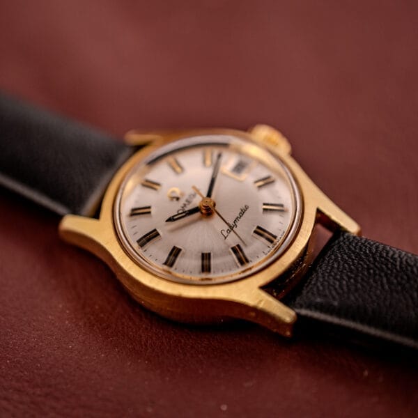 Close-up of an Omega Ladymatic Sunburst Dial Ref. MD 566.021 Cal. 680 gold wristwatch with a black leather strap, resting on a brown surface.