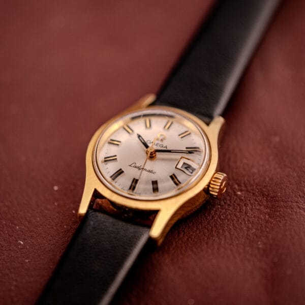 An Omega Ladymatic Sunburst Dial Ref. MD 566.021 Cal. 680 gold wristwatch with a sunburst dial and black leather strap is displayed on a brown textured surface.