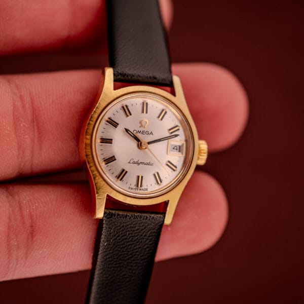 Close-up of a hand holding the Omega Ladymatic Sunburst Dial Ref. MD 566.021 Cal. 680, showcasing its gold case, black leather strap, and white face.