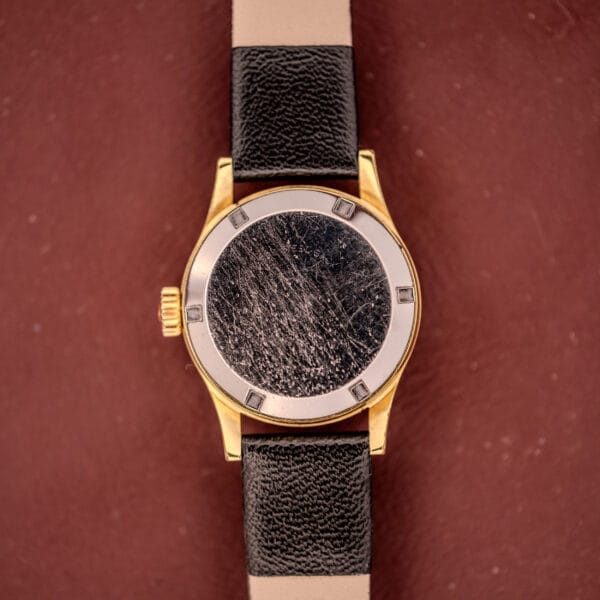 Omega Ladymatic Sunburst Dial Ref. MD 566.021 Cal. 680 gold wristwatch with a heavily scratched, blank sunburst dial and black leather strap, displayed on a brown background.