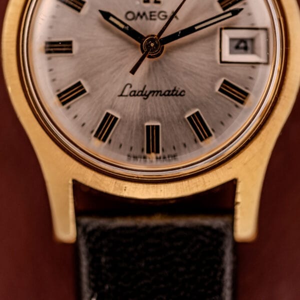 Close-up of an Omega Ladymatic Sunburst Dial Ref. MD 566.021 with Cal. 680 movement, displaying time and date on a gold case paired with a black leather strap.