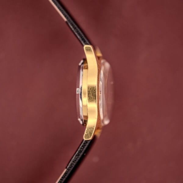 Side view of the Omega Ladymatic Sunburst Dial Ref. MD 566.021 Cal. 680, featuring a gold case, black leather strap, and sunburst dial, set against a blurred maroon background.