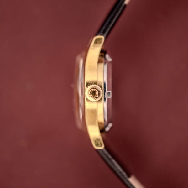 Close-up of the Omega Ladymatic Sunburst Dial Ref. MD 566.021 Cal. 680’s gold crown and black leather strap, with its signature sunburst dial, set against a reddish-brown background.
