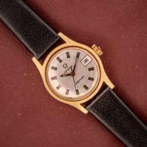 Omega Ladymatic Sunburst Dial Ref. MD 566.021 Cal. 680 gold wristwatch with black leather strap, date display, and sunburst dial shown on a brown background.
