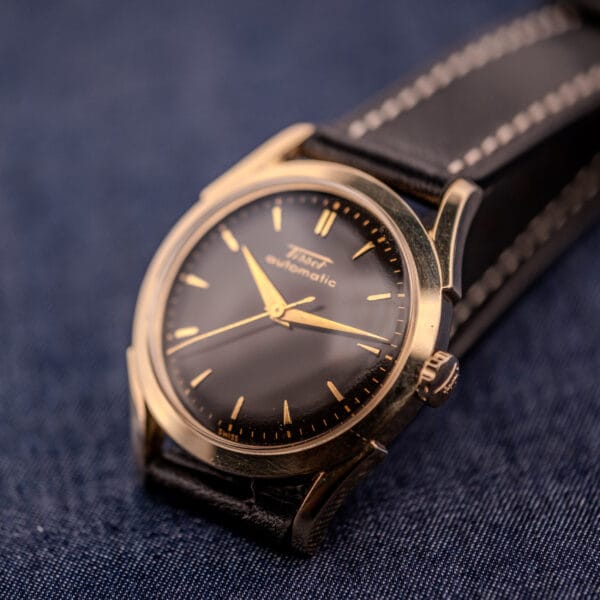 A Tissot Automatic Bumper with a black dial and gold filled case (Cal. 28.5T) rests on dark blue fabric, its luxe finish contrasting with the setting, paired with a black leather strap.