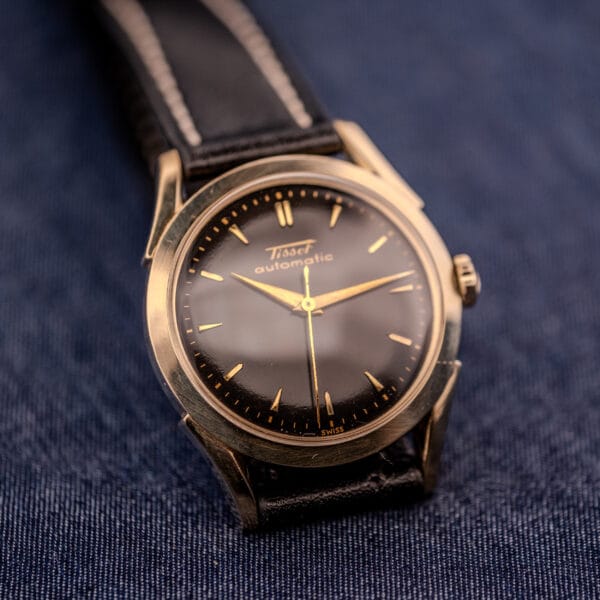 The Tissot Automatic Bumper (Cal. 28.5T) features a black dial, gold filled case, and is paired with a leather strap on dark fabric.