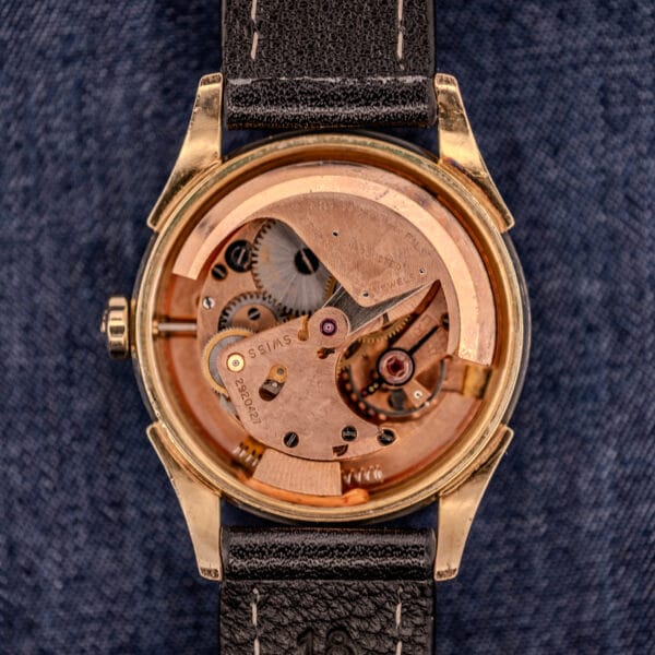 The Tissot Automatic Bumper features a black dial and gold-filled case (Cal. 28.5T), showcasing visible mechanical movement and gears on a dark textured fabric background.