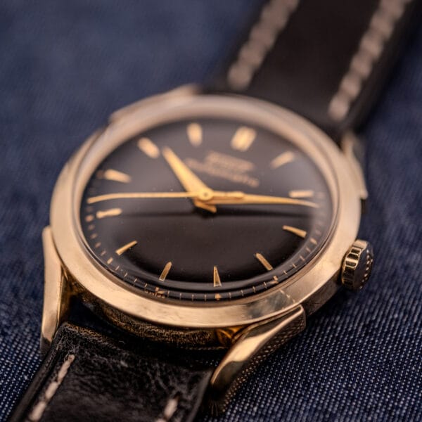 Close-up of a Tissot Automatic Bumper watch, featuring a black dial and gold-filled case (Cal. 28.5T), with a leather strap, set against a blue fabric background.