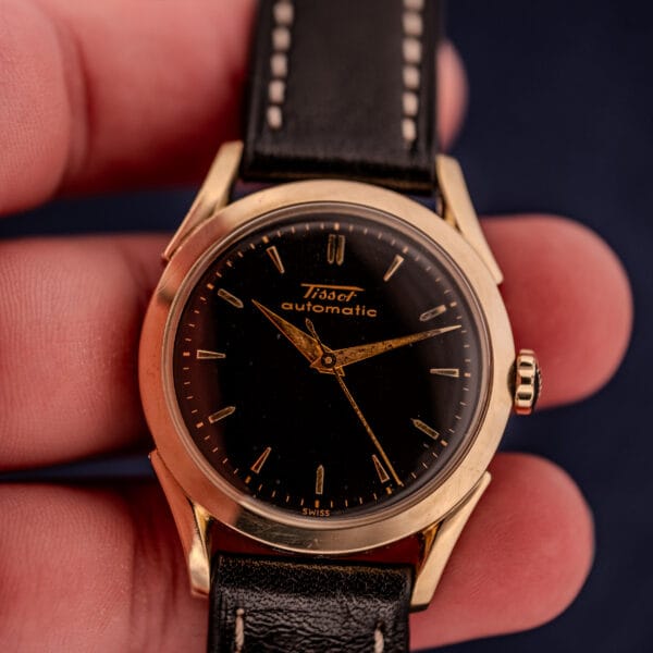A hand holds a Tissot Automatic Bumper wristwatch with a black dial, black leather strap, and gold filled case (Cal. 28.5T).
