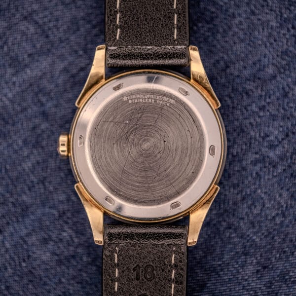 The Tissot Automatic Bumper Black Dial & Gold Filled Case Cal. 28.5T watch with a black leather strap rests on blue textured fabric, showing its elegant gold filled case and automatic design.