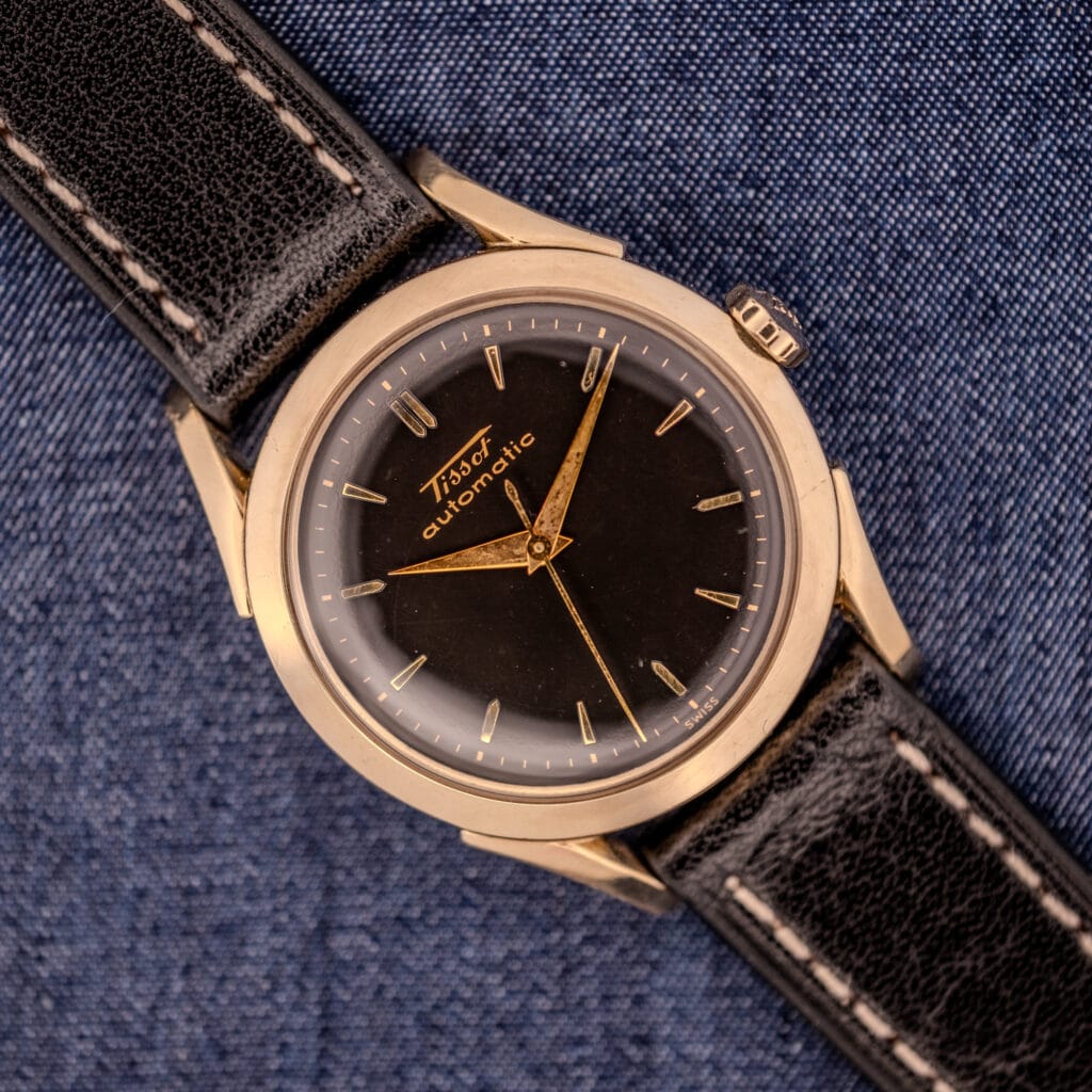 A vintage Tissot Automatic Bumper wristwatch, featuring a black dial and gold filled case (Cal. 28.5T), is displayed elegantly atop blue denim fabric.