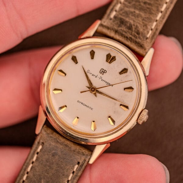 A hand holds a vintage Girard Perregaux Gyromatic Ref. B-1852 wristwatch, showcasing a gold case, brown leather strap, and the renowned Automatic Cal. 21 movement.