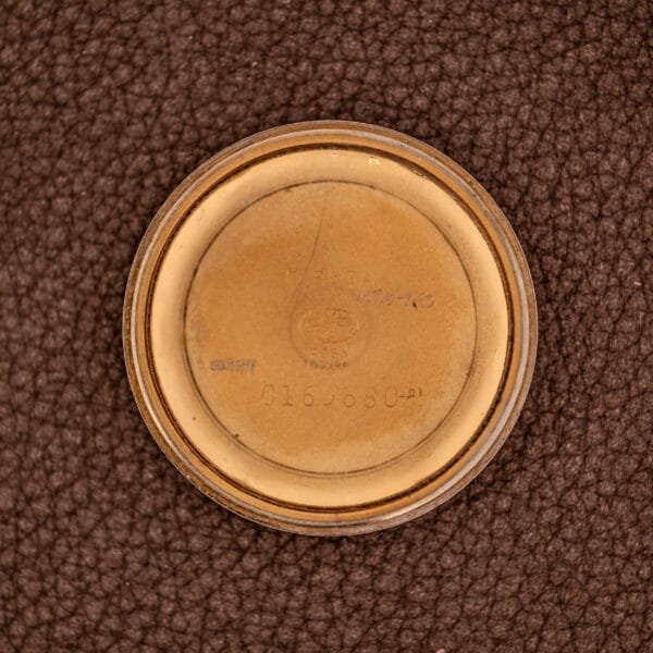 Round beige container lid with faint numbers and text, placed on a textured brown surface—possibly associated with a Girard Perregaux Gyromatic Ref. B-1852 Automatic Cal. 21 watch component.