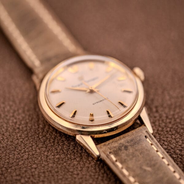 A Girard Perregaux Gyromatic Ref. B-1852 gold wristwatch with a leather strap and Automatic Cal. 21 rests on a textured brown surface.