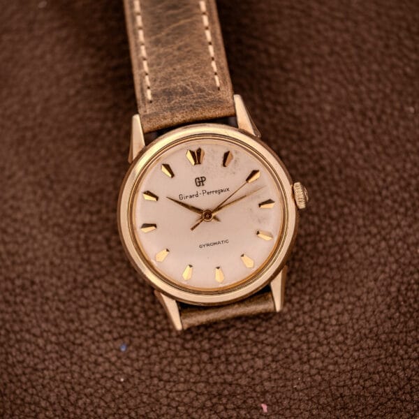 A Girard Perregaux Gyromatic Ref. B-1852 Automatic Cal. 21 wristwatch with a brown leather strap rests on a textured brown surface.