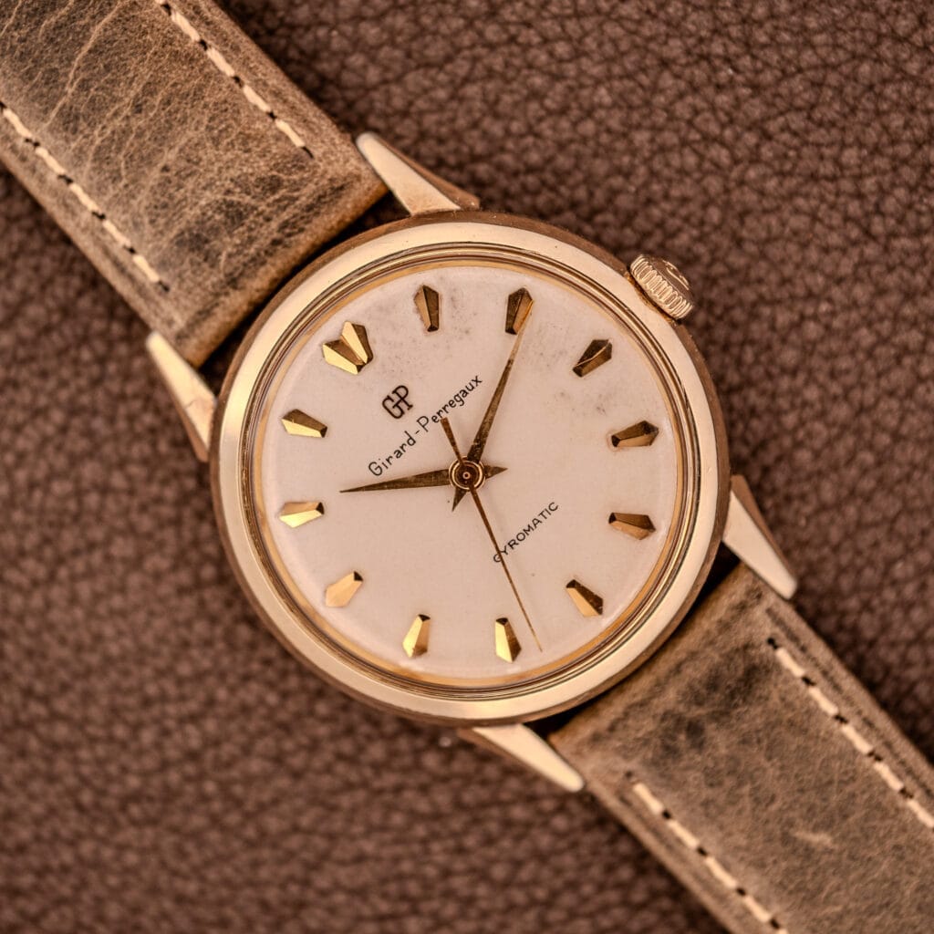 A Girard Perregaux Gyromatic Ref. B-1852 Automatic Cal. 21 wristwatch with a gold case and brown leather strap rests elegantly on a textured surface.