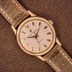 A Girard Perregaux Gyromatic Ref. B-1852 Automatic Cal. 21 wristwatch with a gold case and brown leather strap rests elegantly on a textured surface.