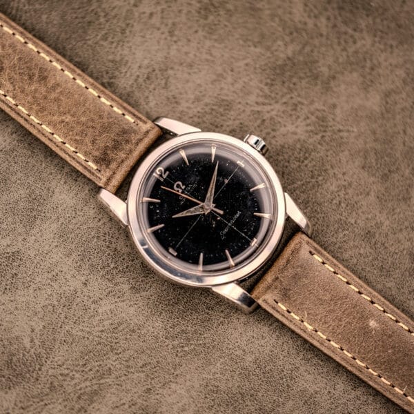 An Omega Automatic Seamaster Ref. 2848 Black Dial Cal. 500 wristwatch with a black dial and tan leather strap rests on a textured brown surface.