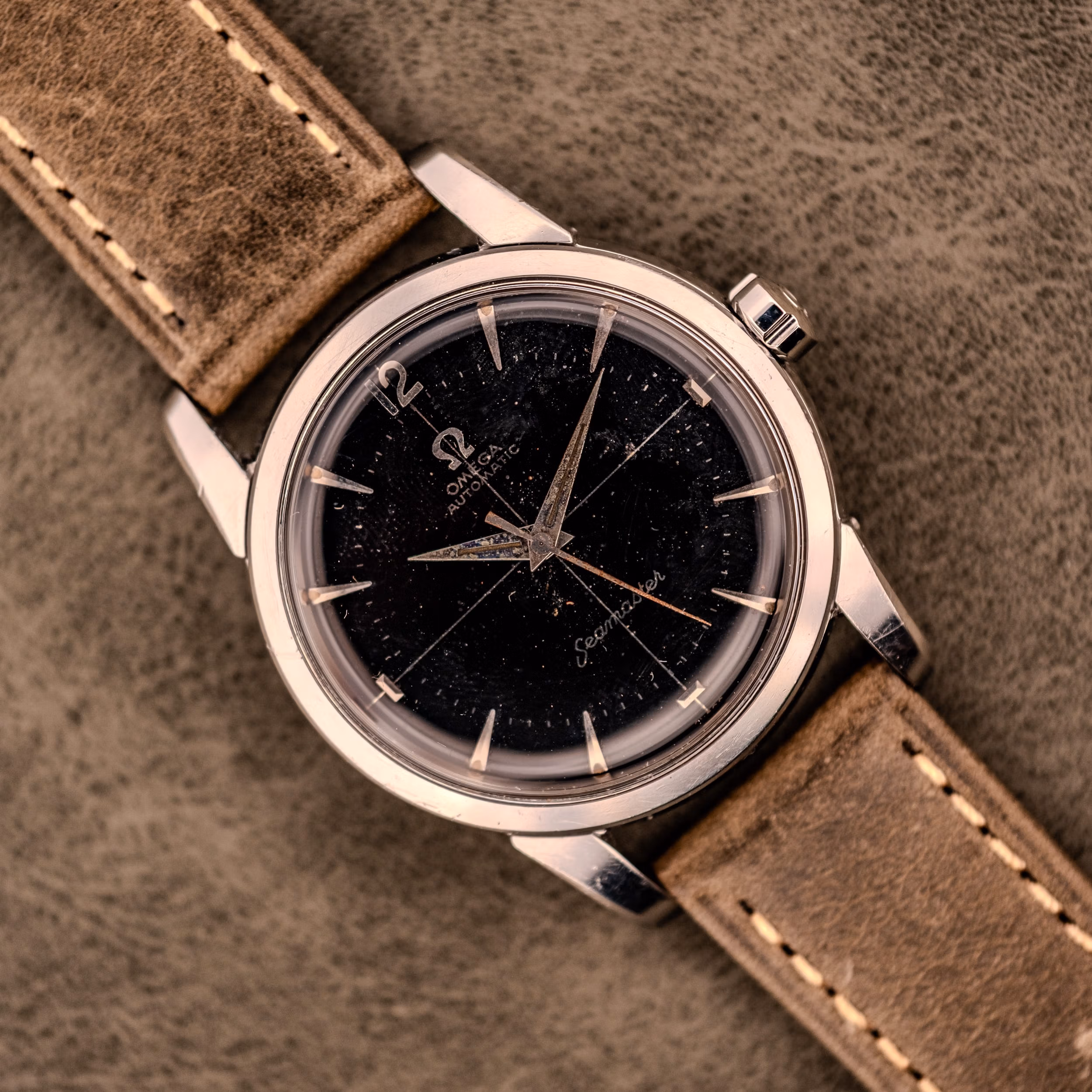 An Omega Automatic Seamaster Ref. 2848 with a black dial and brown leather strap, Cal. 500, displayed on a suede surface. Omega Seamaster Ref. 2848 with original black dial showing speckled patina and applied silver dagger indices on olive leather