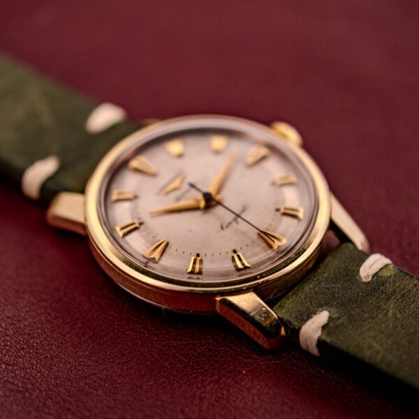 Longines Automatic Conquest Ref. 9000-1 14k Gold Capped Case wristwatch with green leather strap on a burgundy surface, featuring Roman numeral markers.