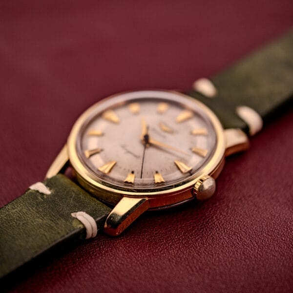 Longines Automatic Conquest Ref. 9000-1 wristwatch with a 14k gold capped case and green leather strap, displayed on a textured burgundy surface.