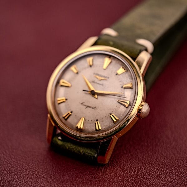 The Longines Automatic Conquest Ref. 9000-1, featuring a 14k gold capped case and green leather strap, is elegantly displayed on a red textured surface.