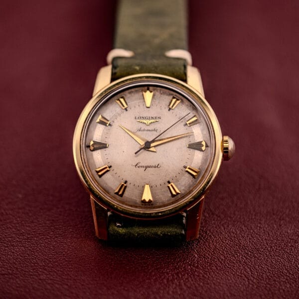 A Longines Automatic Conquest Ref. 9000-1 with a 14k gold capped case and green leather strap is elegantly displayed on a maroon surface.