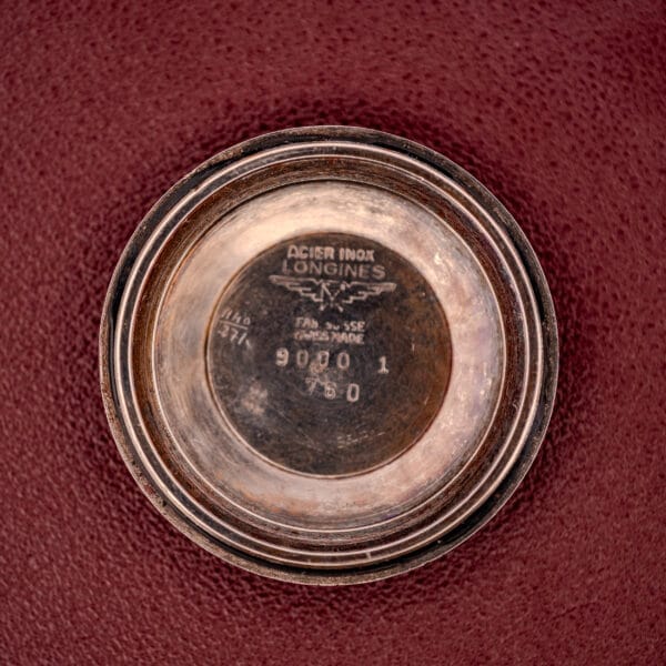 Round metal case back engraved with the Longines logo, Ref. 9000-1, and serial numbers—shown on a textured red surface—highlights the refined details of the Longines Automatic Conquest Ref. 9000-1 14k Gold Capped Case.