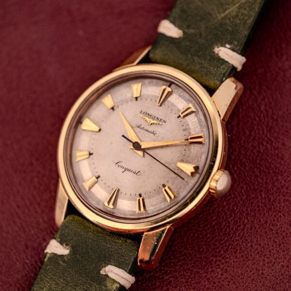 A Longines Automatic Conquest Ref. 9000-1 watch with a 14k gold capped case and green leather strap, displayed on a maroon background.